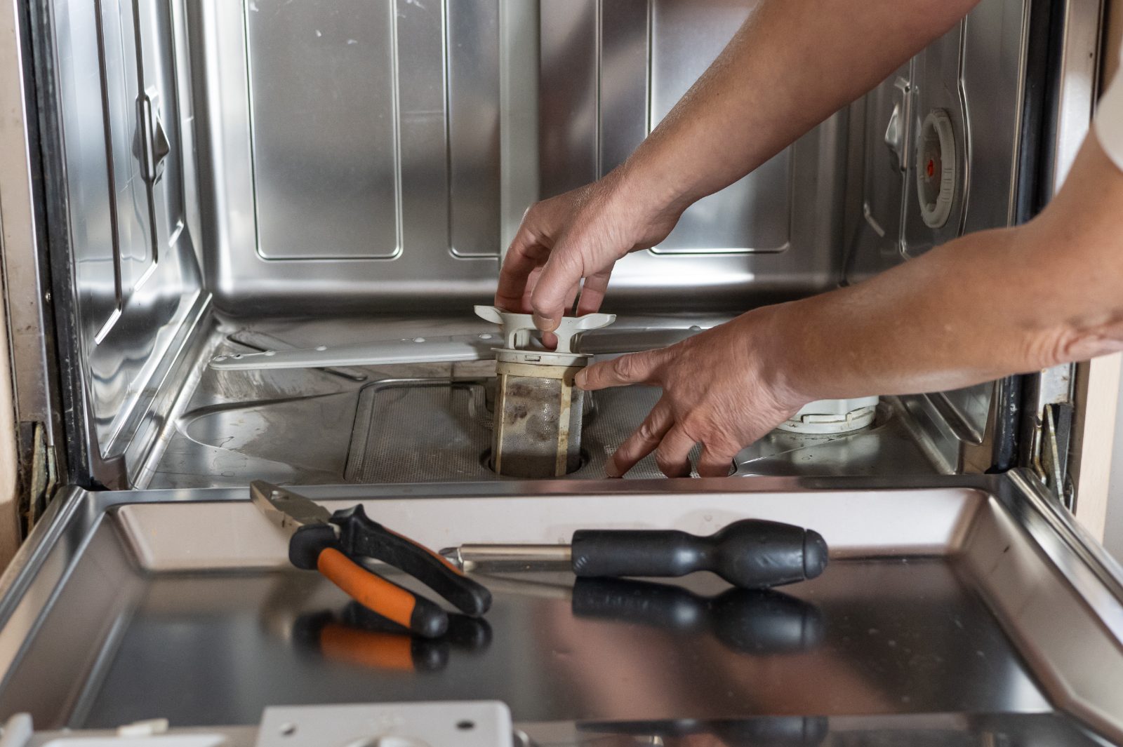 Person repairing dishwasher filter with tools on stainless steel tub